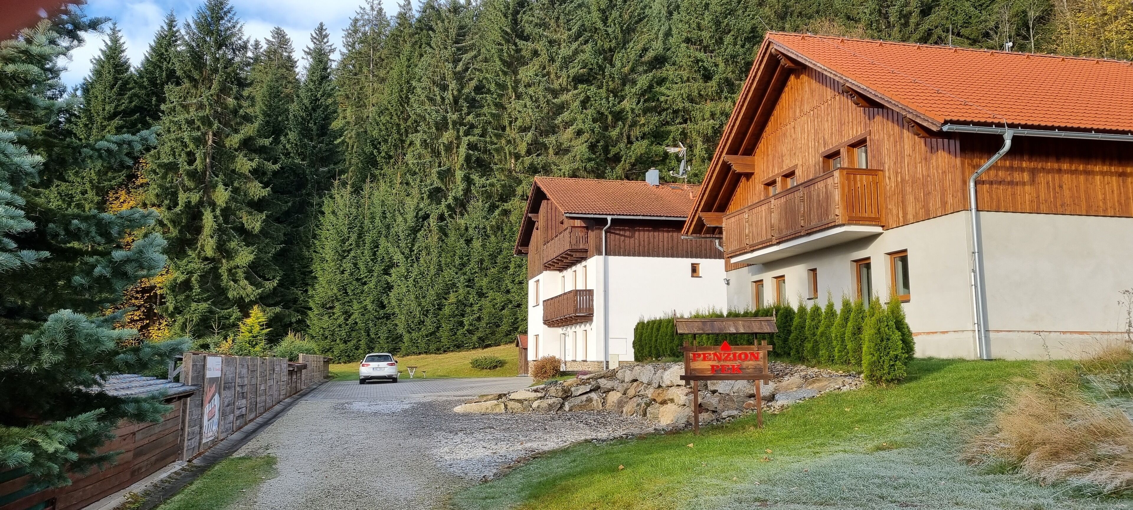 A wooden building with a red-tiled roof, a sign reading "Pension Park," and a gravel driveway leading to a parked car.