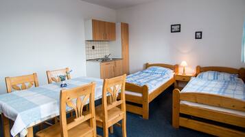 A compact room with two single beds, a wooden dining table with chairs, a small kitchenette, and a nightstand with a lamp.