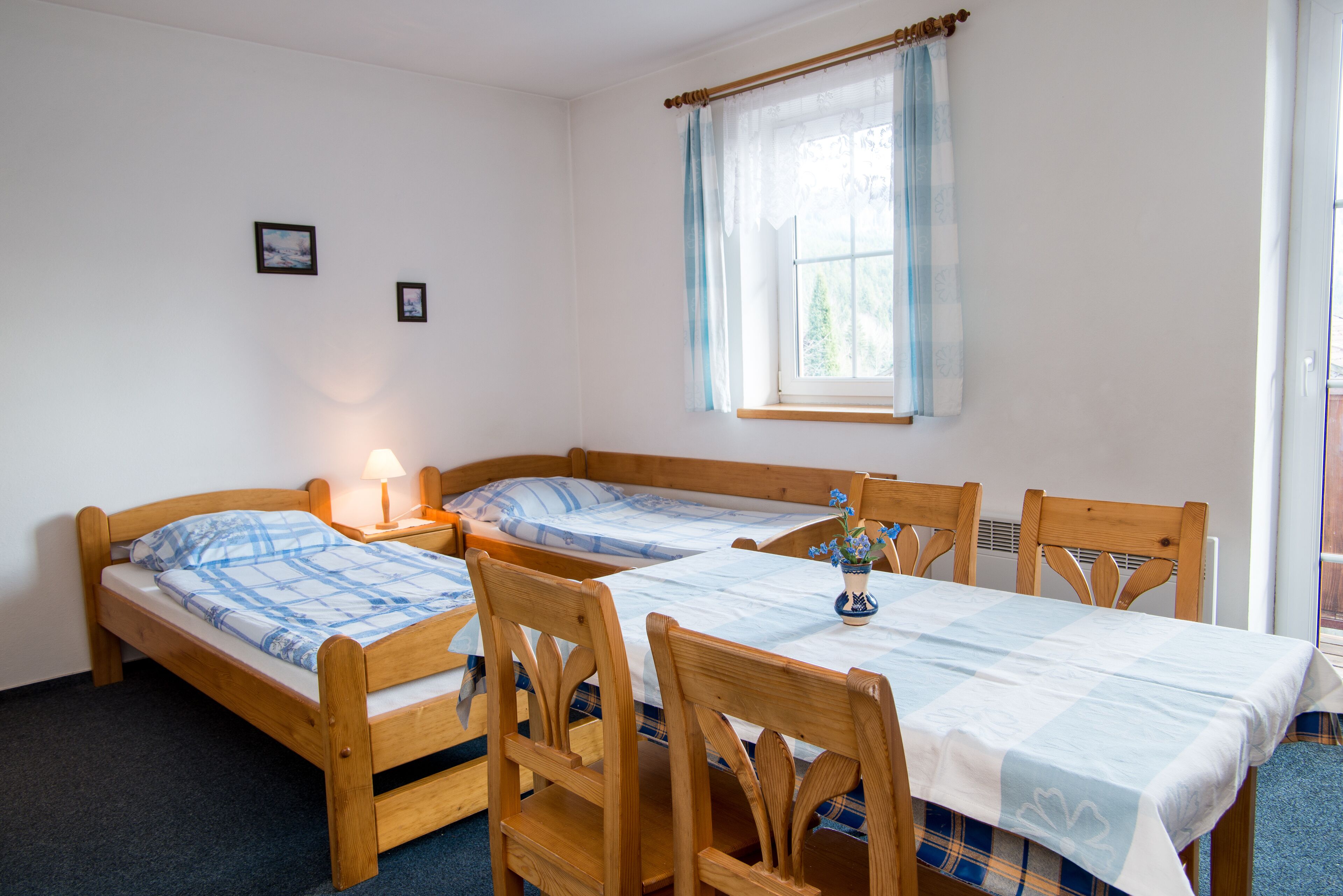 A room with two wooden beds, a dining table with chairs, a window with a curtain, and a small table with a lamp.