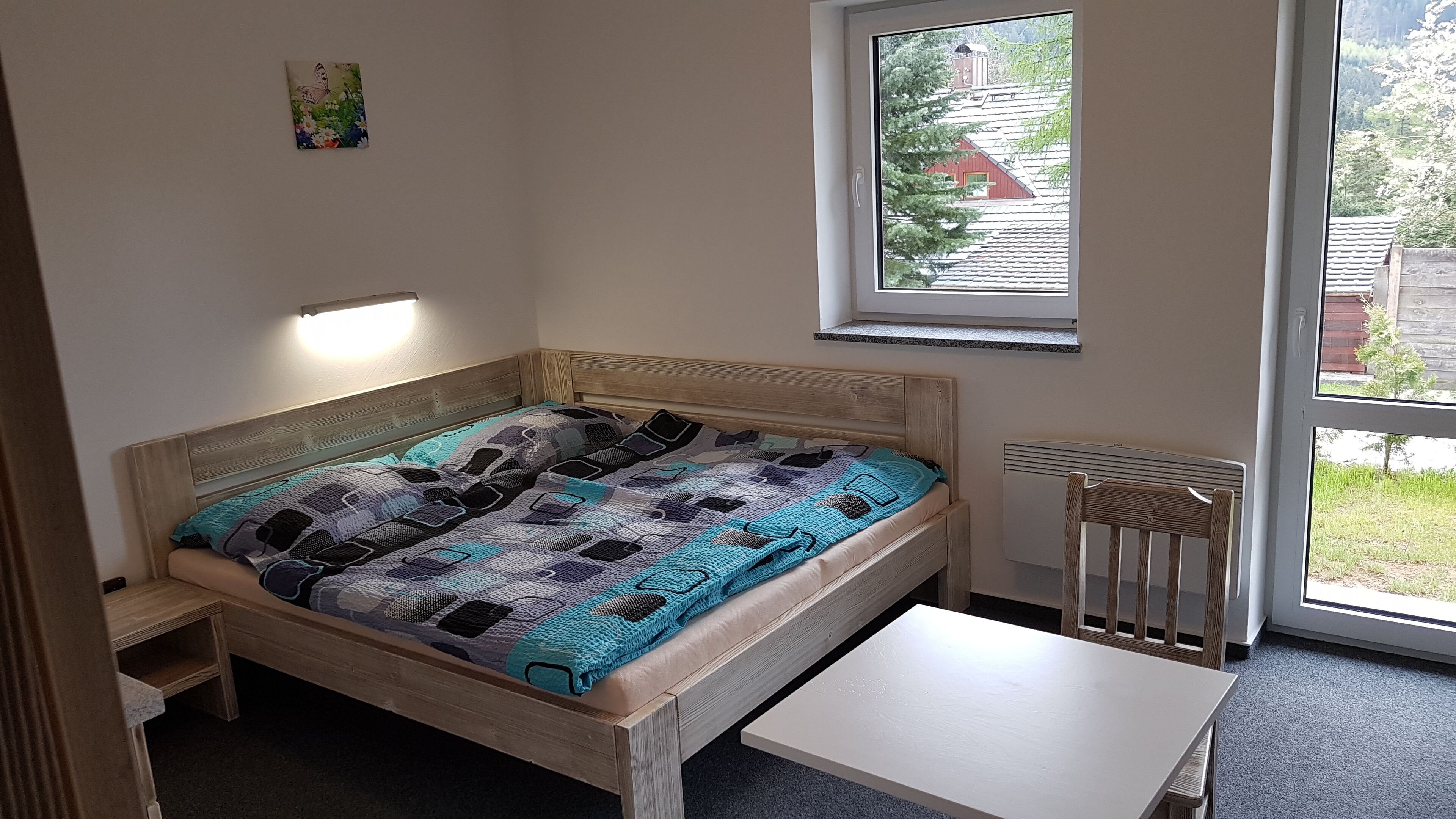 Un letto singolo con coperta decorata, un comodino, un termosifone e una finestra con vista su una casa e degli alberi.