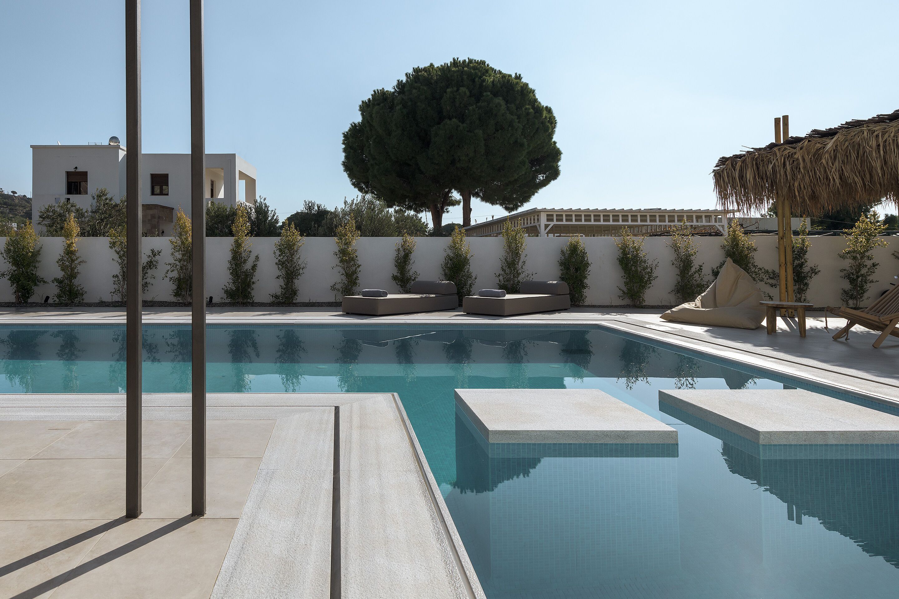 A swimming pool with clear blue water, surrounded by a low white wall, and a large tree in the background.