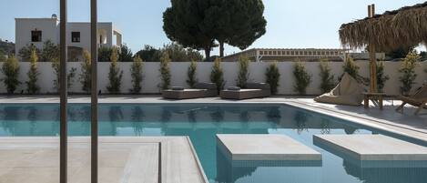 A swimming pool with clear blue water, surrounded by a low white wall, and a large tree in the background.