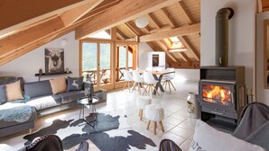 Ein großzügiger Loft mit hohen Decken, Holzbalken, einem Kamin, einer Couch, einem Esstisch und Blick ins Freie.