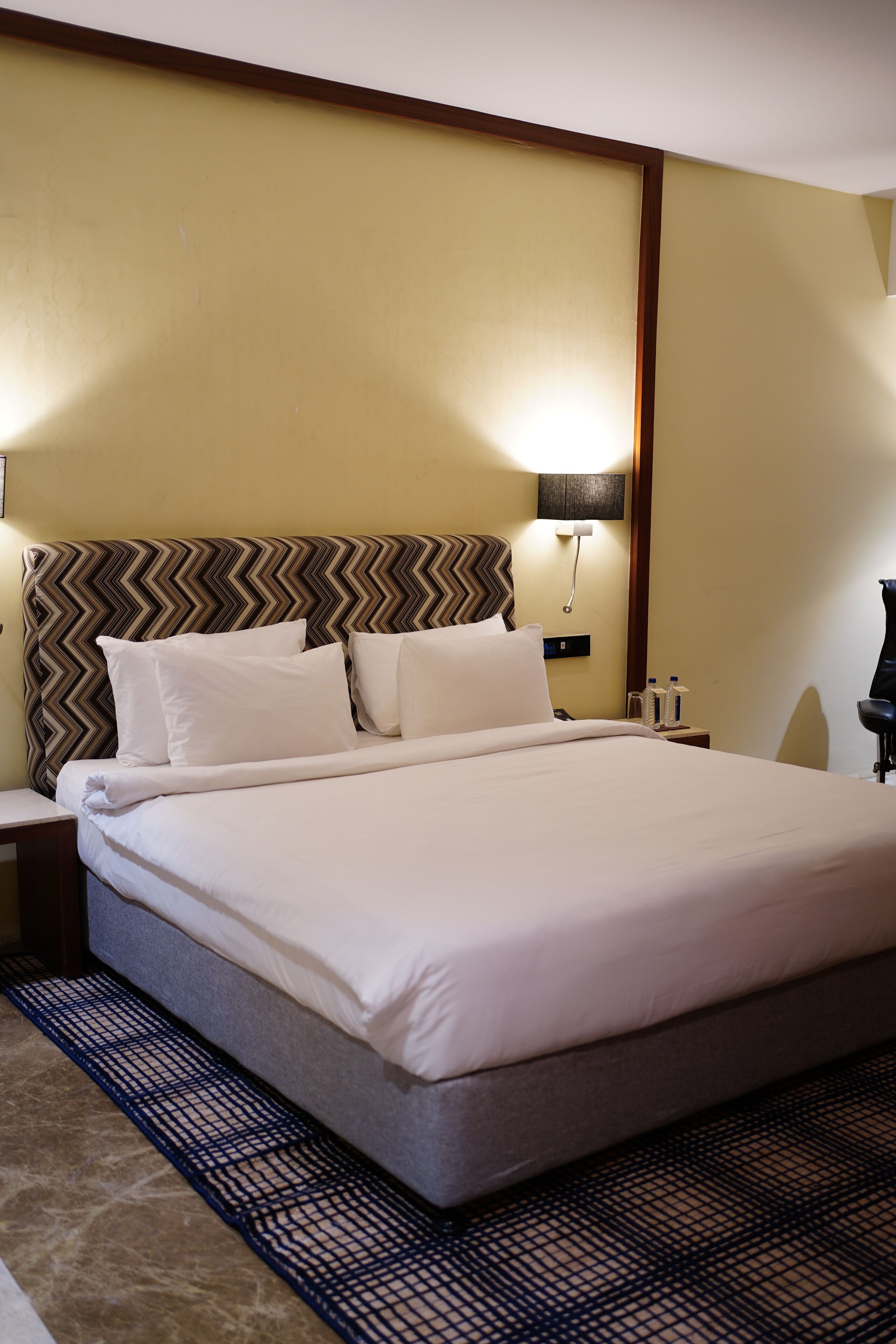 A hotel room with a large bed, a patterned headboard, a nightstand, a lamp, and a wall-mounted phone.