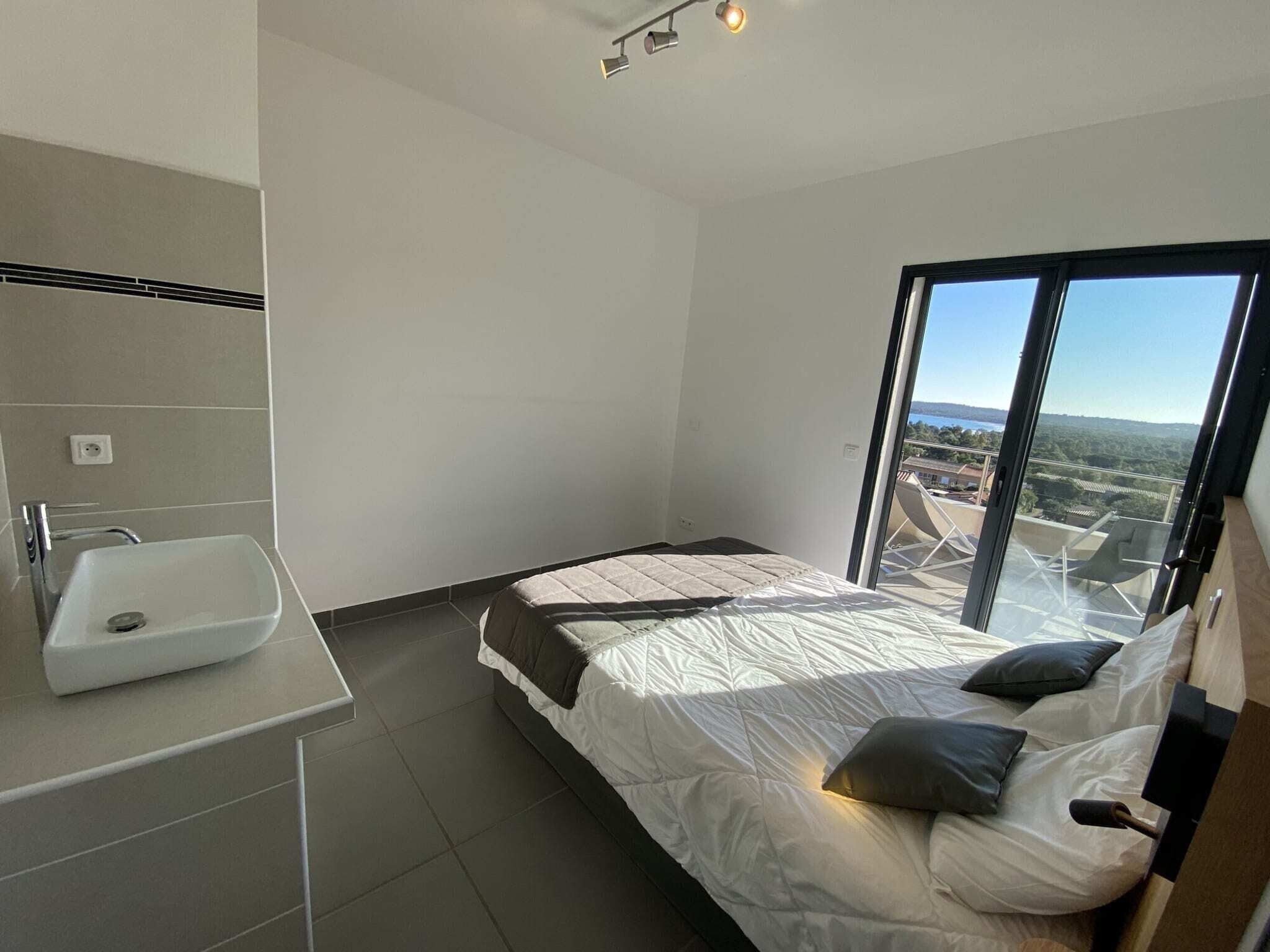 A modern bedroom with a large bed, a bedside table, a built-in sink, and a large window offering a view of the landscape.