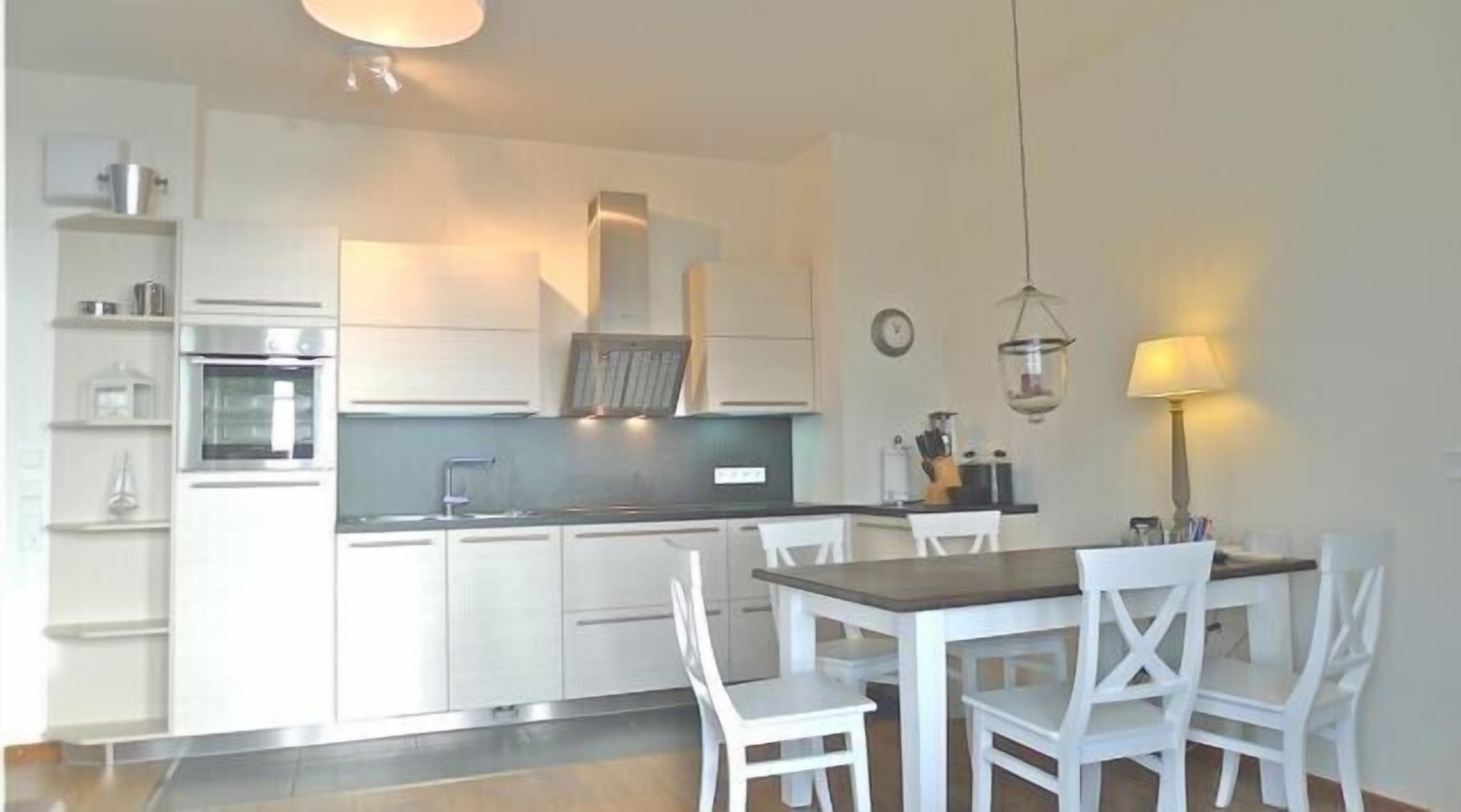 Une cuisine moderne avec des meubles blancs, un four encastré et un coin repas comprenant une table et des chaises.