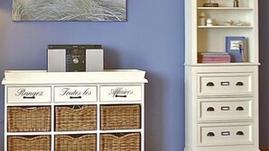 Une pièce avec une armoire blanche et des paniers en osier étiquetés « Rangée », « Toutes » et « Affaires ». Une bibliothèque blanche avec le mot « Armadillo » écrit sur l’étagère supérieure et divers objets sur les étagères inférieures. Un tableau représentant un phare est accroché au mur.