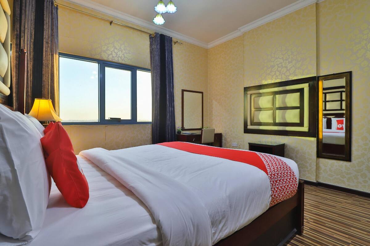 Ein Hotelzimmer mit einem großen Bett, einem Schreibtisch, einem Stuhl, einer Lampe, einem Spiegel und einem Fenster mit Vorhängen.