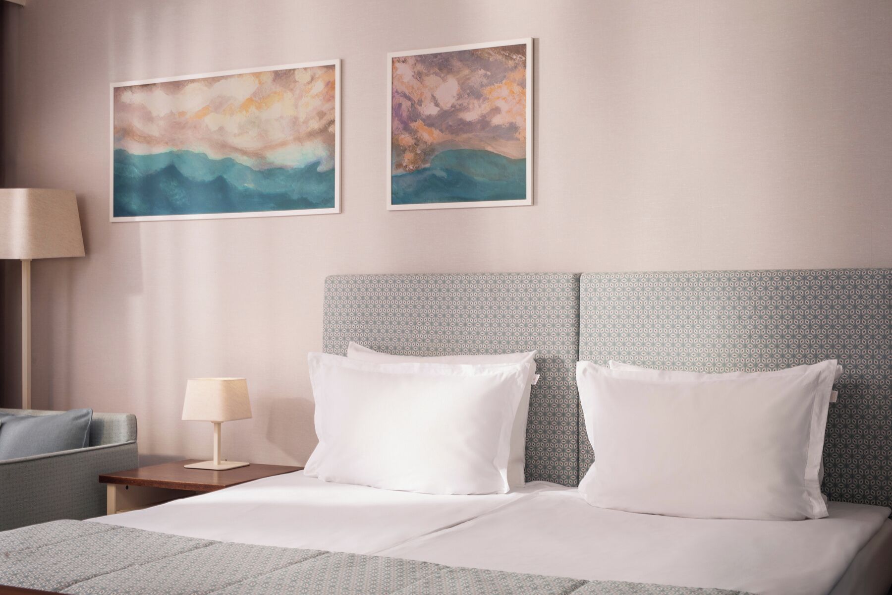 Una habitación de hotel con cama, dos cuadros en la pared, una mesita de noche con lámpara y un cabecero estampado.