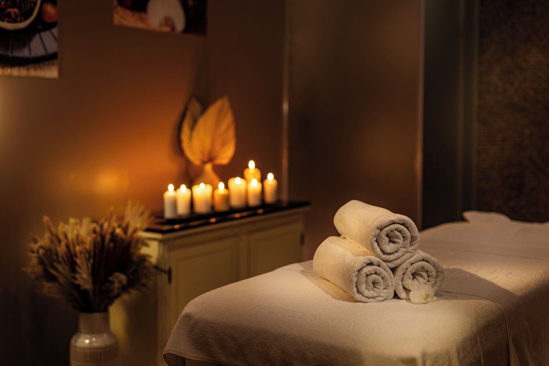 Un cuarto de spa con una camilla de masaje, toallas enrolladas, velas y un florero con flores secas.