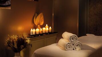 Un cuarto de spa con una camilla de masaje, toallas enrolladas, velas y un florero con flores secas.