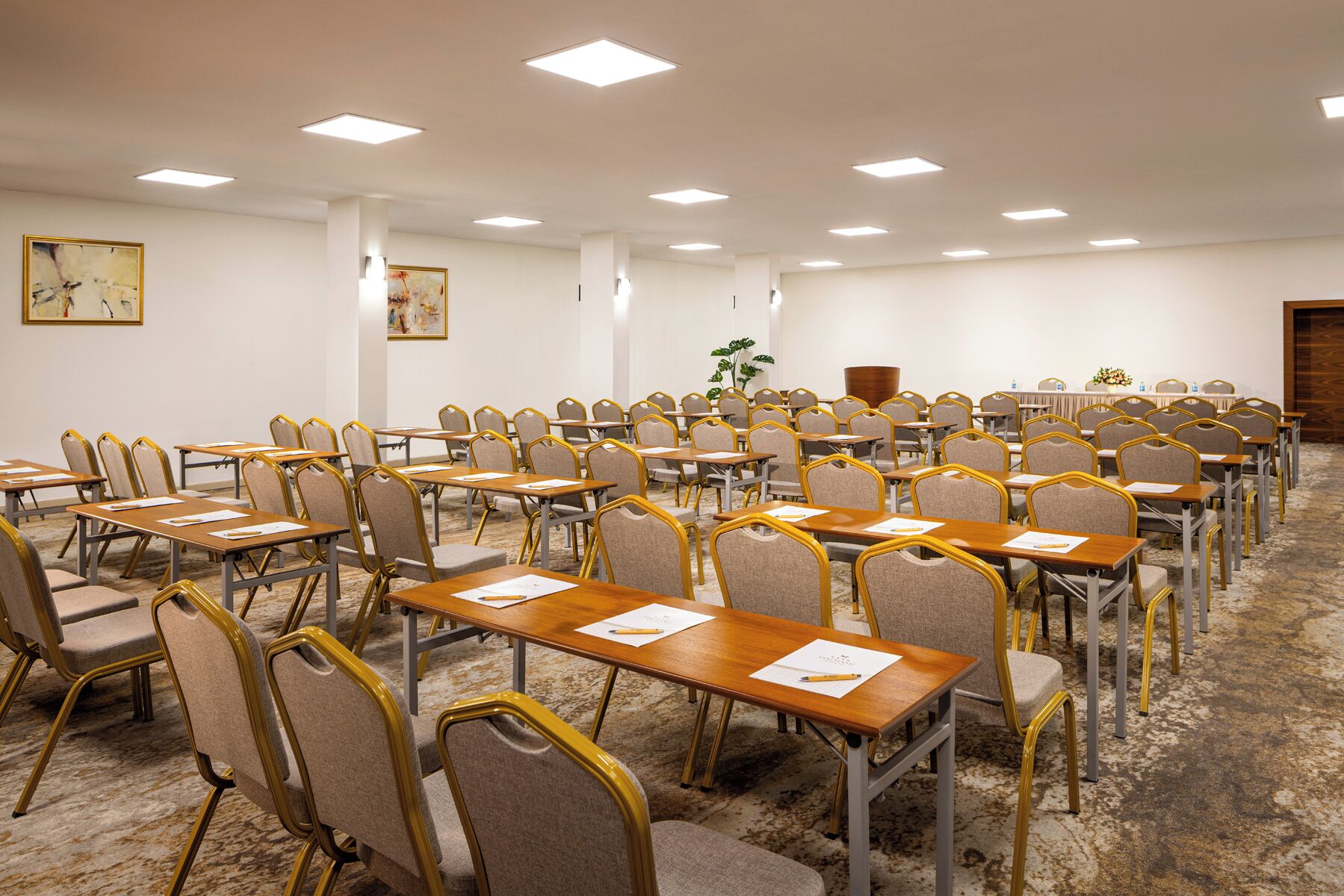 A conference room with rows of tables and chairs, ceiling lights, and a carpeted floor.