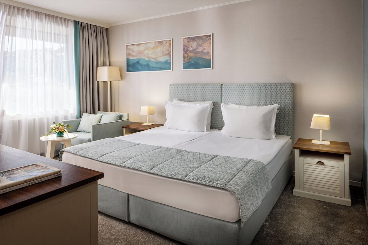 A hotel room with a large bed, two bedside tables, a desk, and a chair. There are two paintings on the wall and a window with curtains.