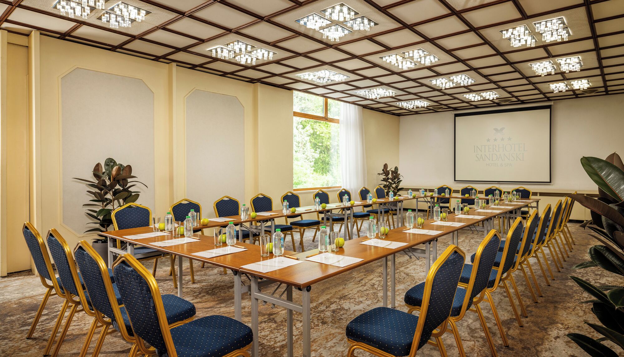 Un salón de conferencias con una larga mesa, sillas, botellas de agua y una pantalla de proyector.