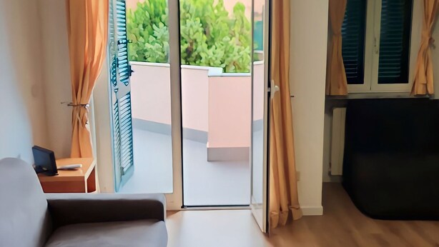 Een kamer met een bank, een klein tafeltje met een boek, een klok en uitzicht op een balkon met planten.