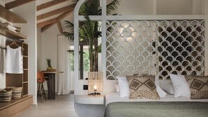 Un dormitorio con cama, cabecera blanca con diseño geométrico, mesita de noche con lámpara, silla de madera y una pequeña mesa con una piña encima.