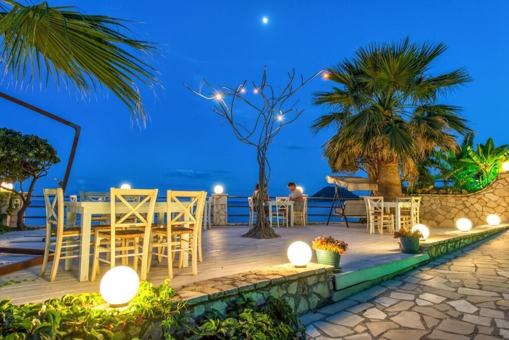 An outdoor dining area with tables and chairs, a tree, and a view of the sea at dusk.