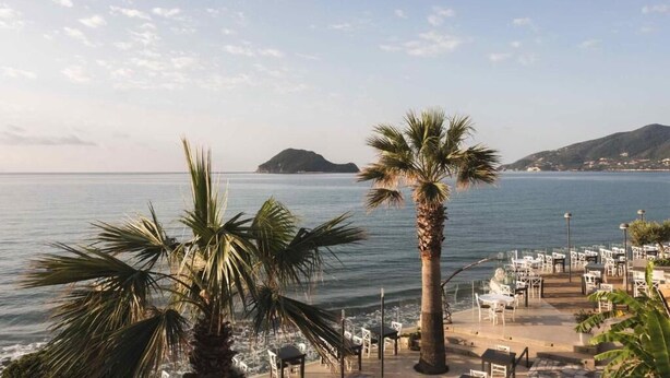 Un restaurante frente al mar con terraza al aire libre, palmeras y colinas a lo lejos.