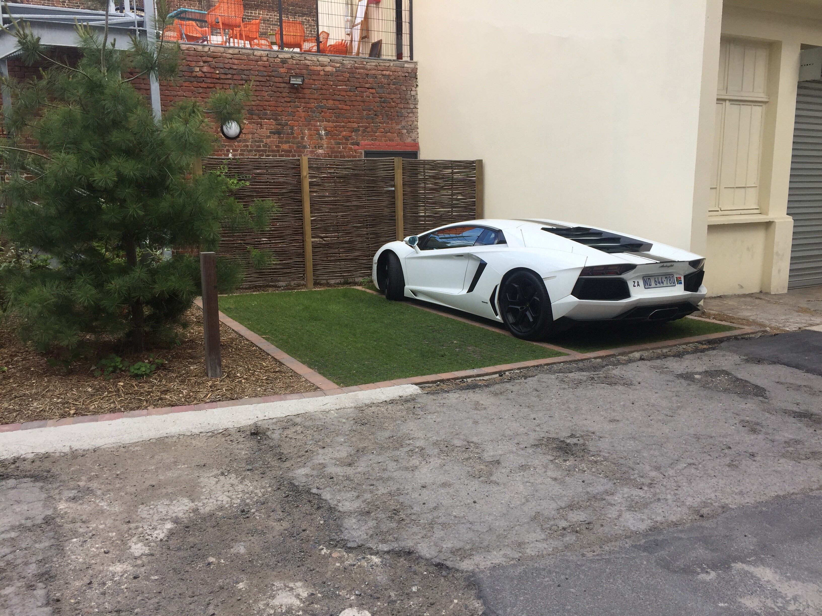 A white sports car parked in a driveway with a small lawn and a brick wall in the background.
