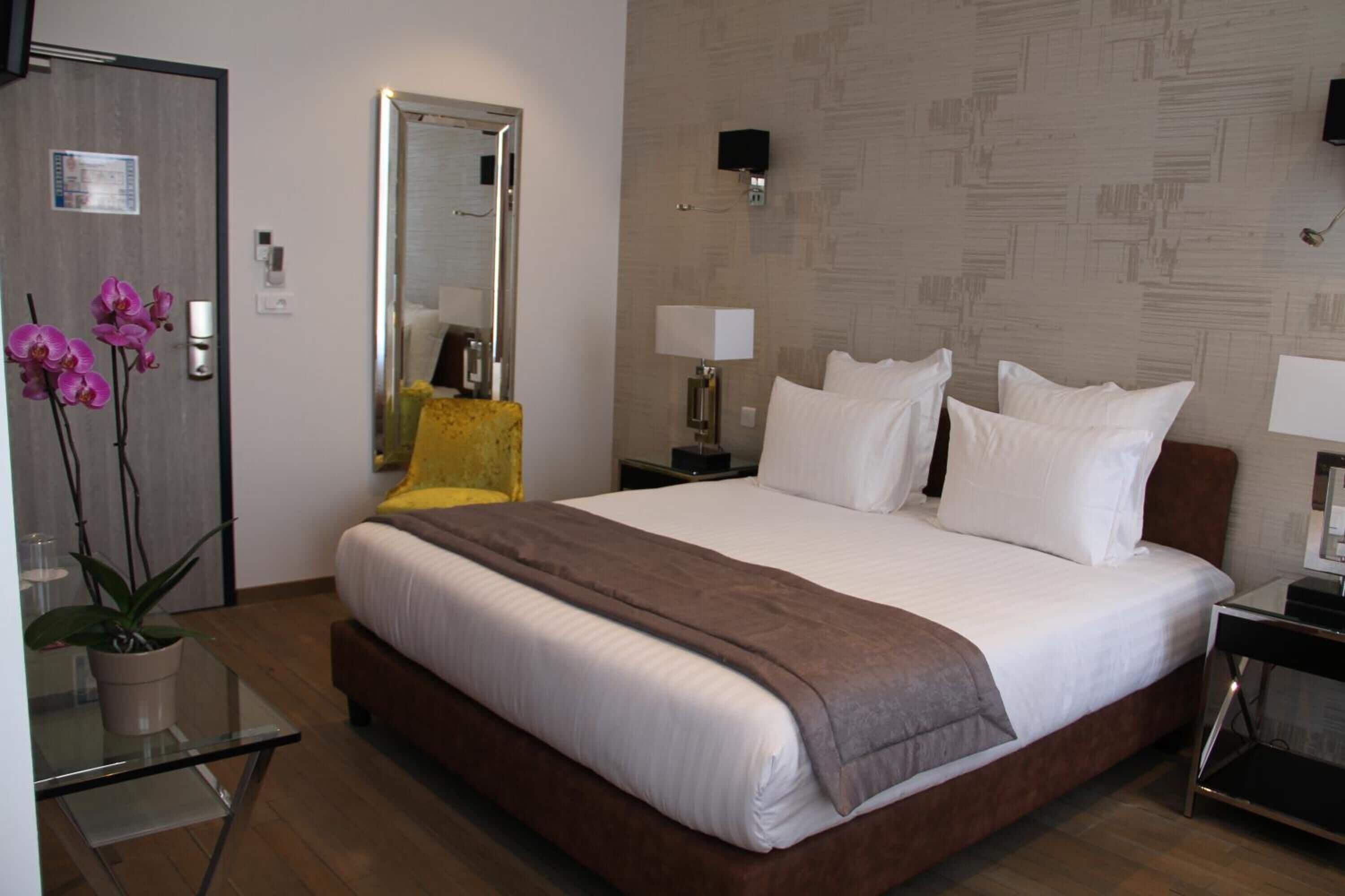 A hotel room with a large bed, bedside lamps, a mirror, a small table with a plant, and a yellow chair.