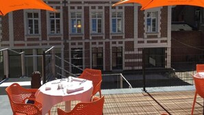 An outdoor seating area with orange umbrellas, a brick building in the background, and a metal railing. - Hotel Le Dome (Arras)