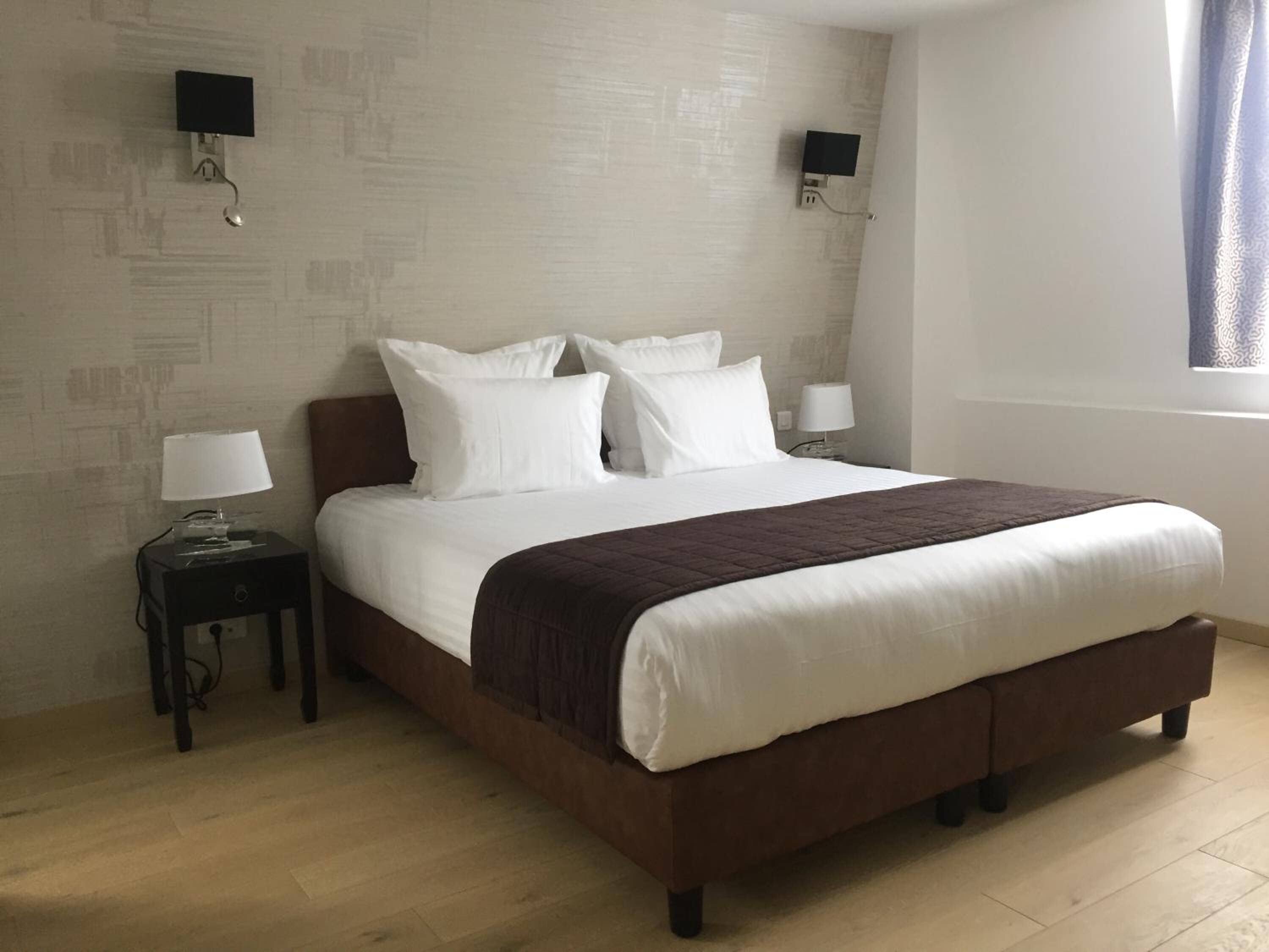 Una habitación de hotel con una cama grande, dos mesitas de noche con lámparas, un televisor montado en la pared y una ventana con cortinas.