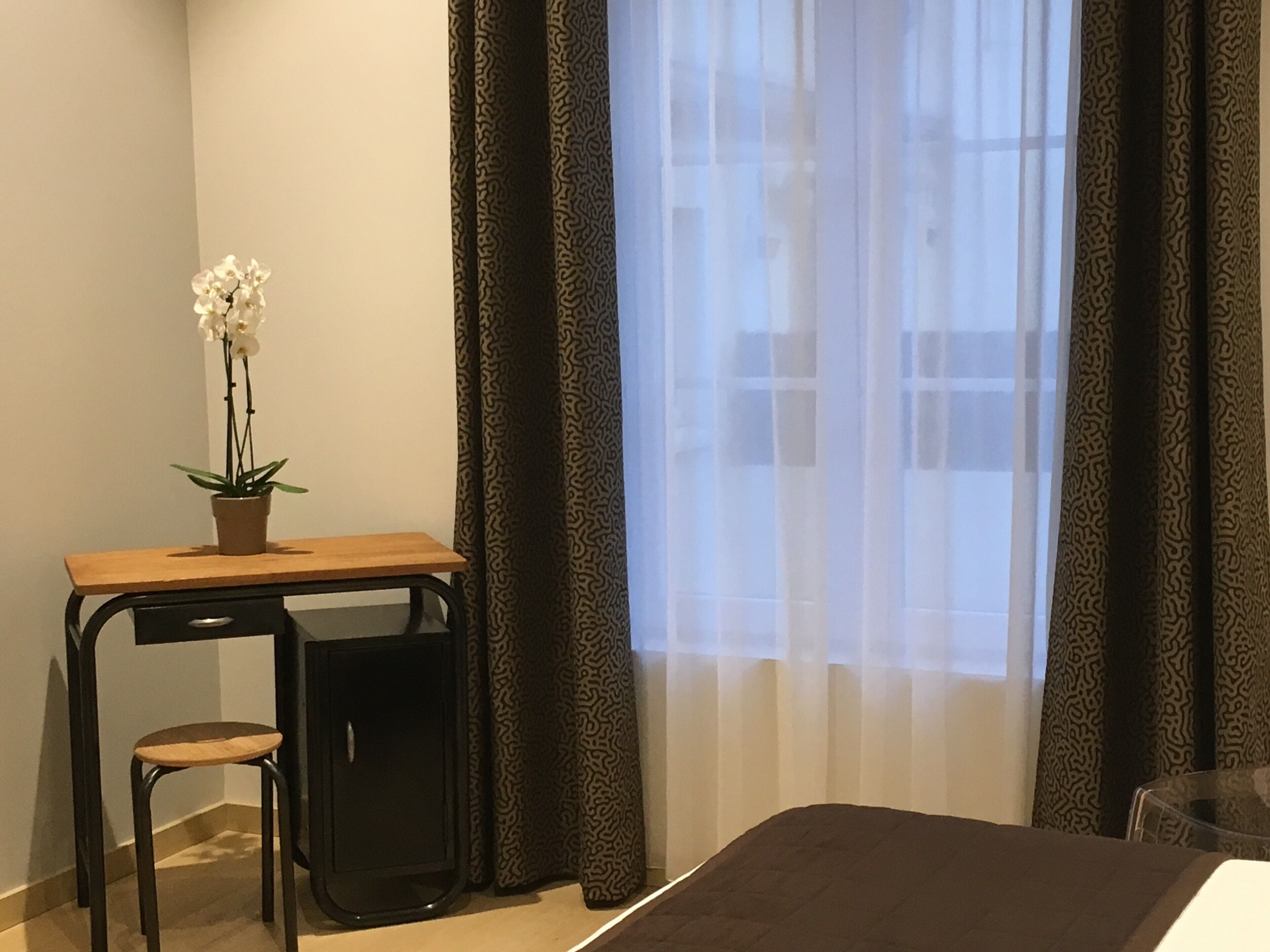 Ein kleiner Raum mit Schreibtisch, Stuhl, Orchidee im Topf und einem Fenster mit gemusterten Vorhängen.