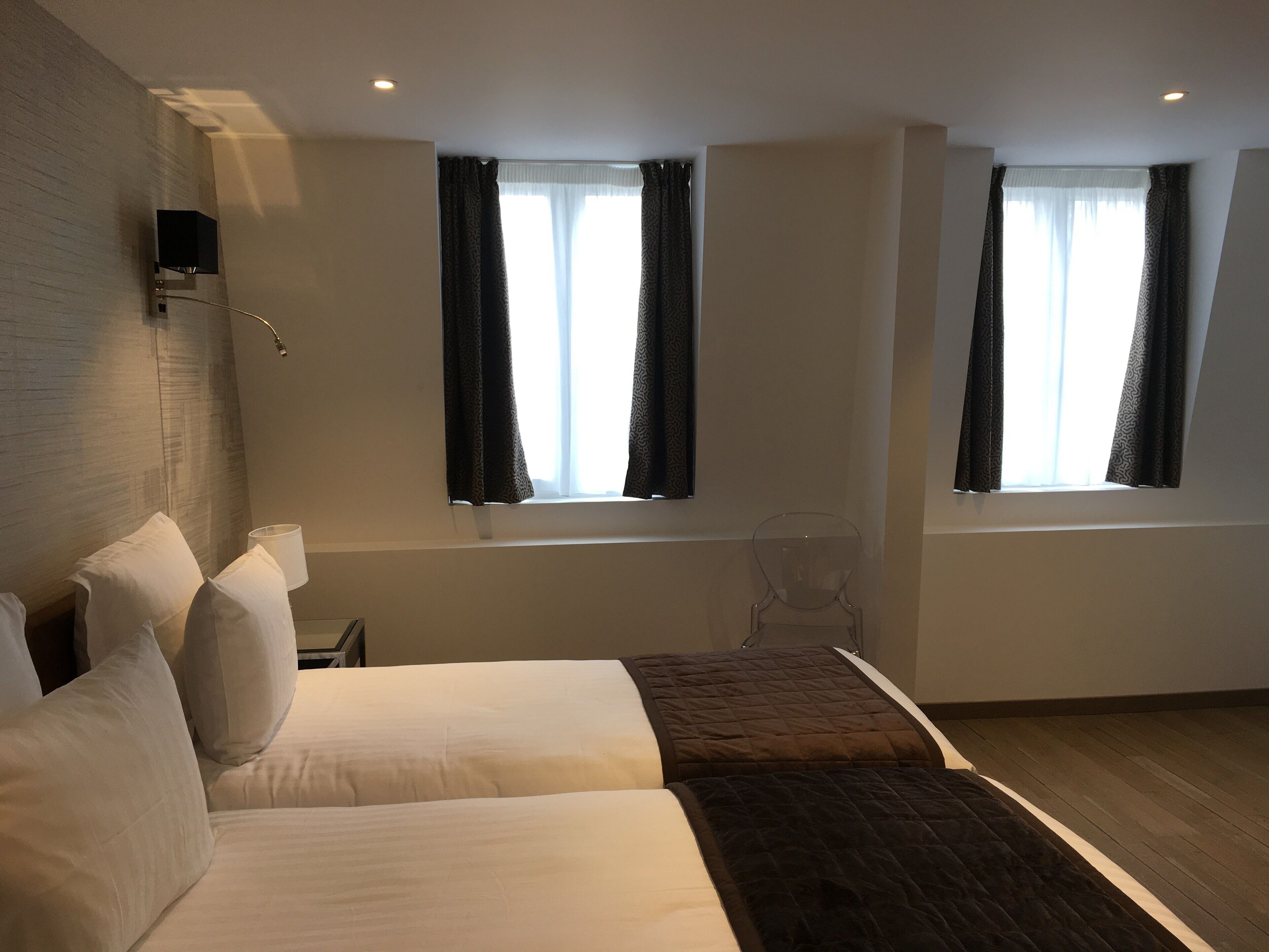 A modern hotel room with two beds, a large window with curtains, a bedside table, and a wall-mounted lamp.