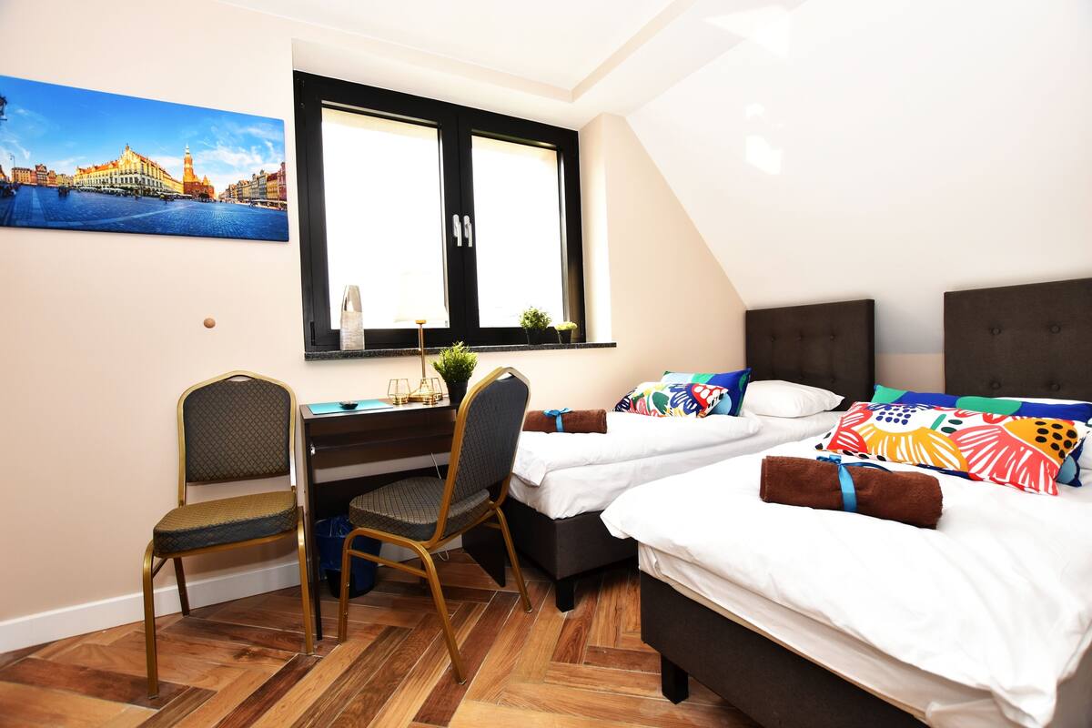 A room with two beds, a desk, and a chair. There is a window with a view of a building.
