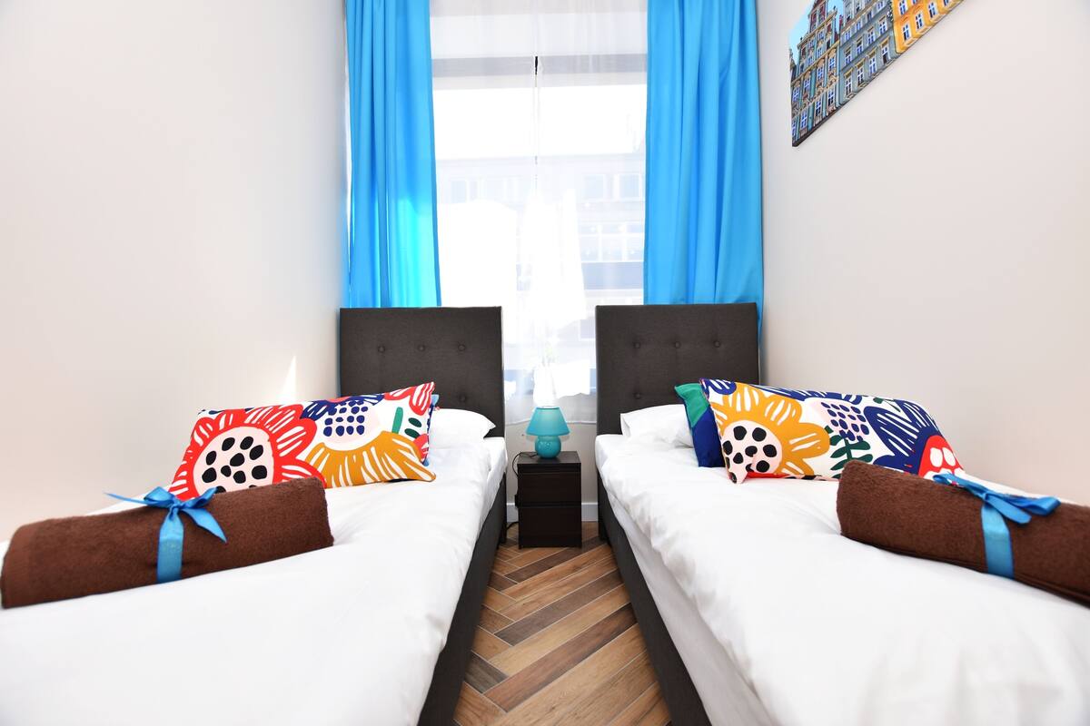A hotel room with two beds, each with colorful patterned pillows and a blue lamp between them.