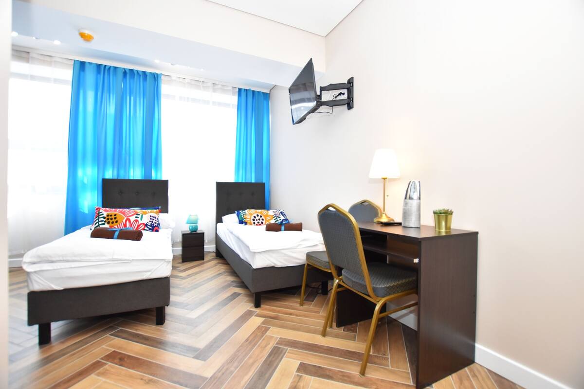 A hotel room with two beds, a desk, a chair, a lamp, and a wall-mounted TV.