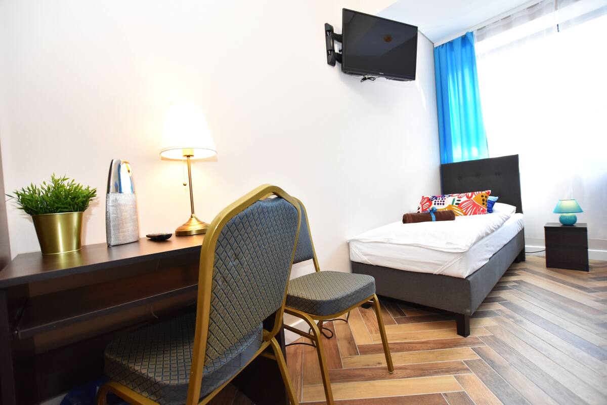 A hotel room with a bed, a desk with a lamp, a chair, a TV mounted on the wall, and a window with blue curtains.