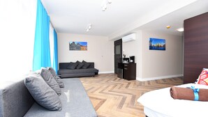 A hotel room with a grey sofa, a bed with a rolled towel, a coffee machine, and two paintings on the walls. - DUKES Apartments - Old Town (Wroclaw)