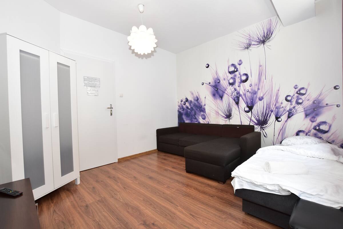 A bedroom with a wooden floor, a black sofa, a bed with white bedding, and a wall mural of purple flowers.
