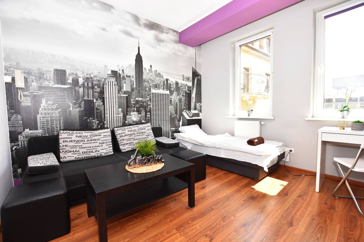 A modern living room with a cityscape mural, a black sofa set, a white bed, and a small desk with a plant.