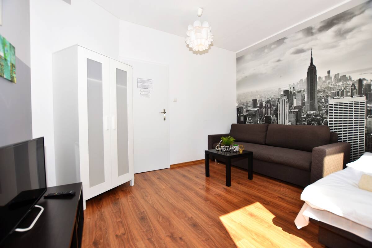 A modern room with a wooden floor, a black sofa, a white wardrobe, a black coffee table, and a cityscape mural on the wall.
