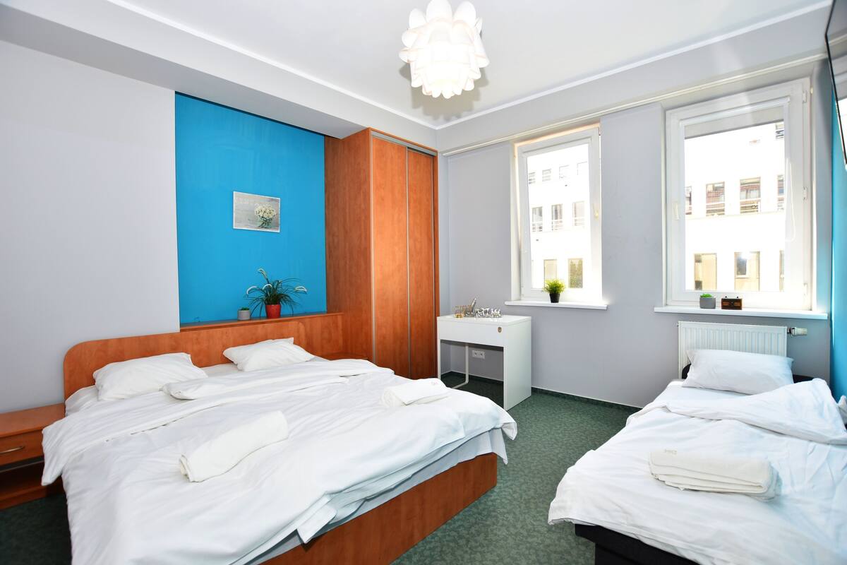 A hotel room with two beds, a desk, a window with a view of buildings, and a blue accent wall.