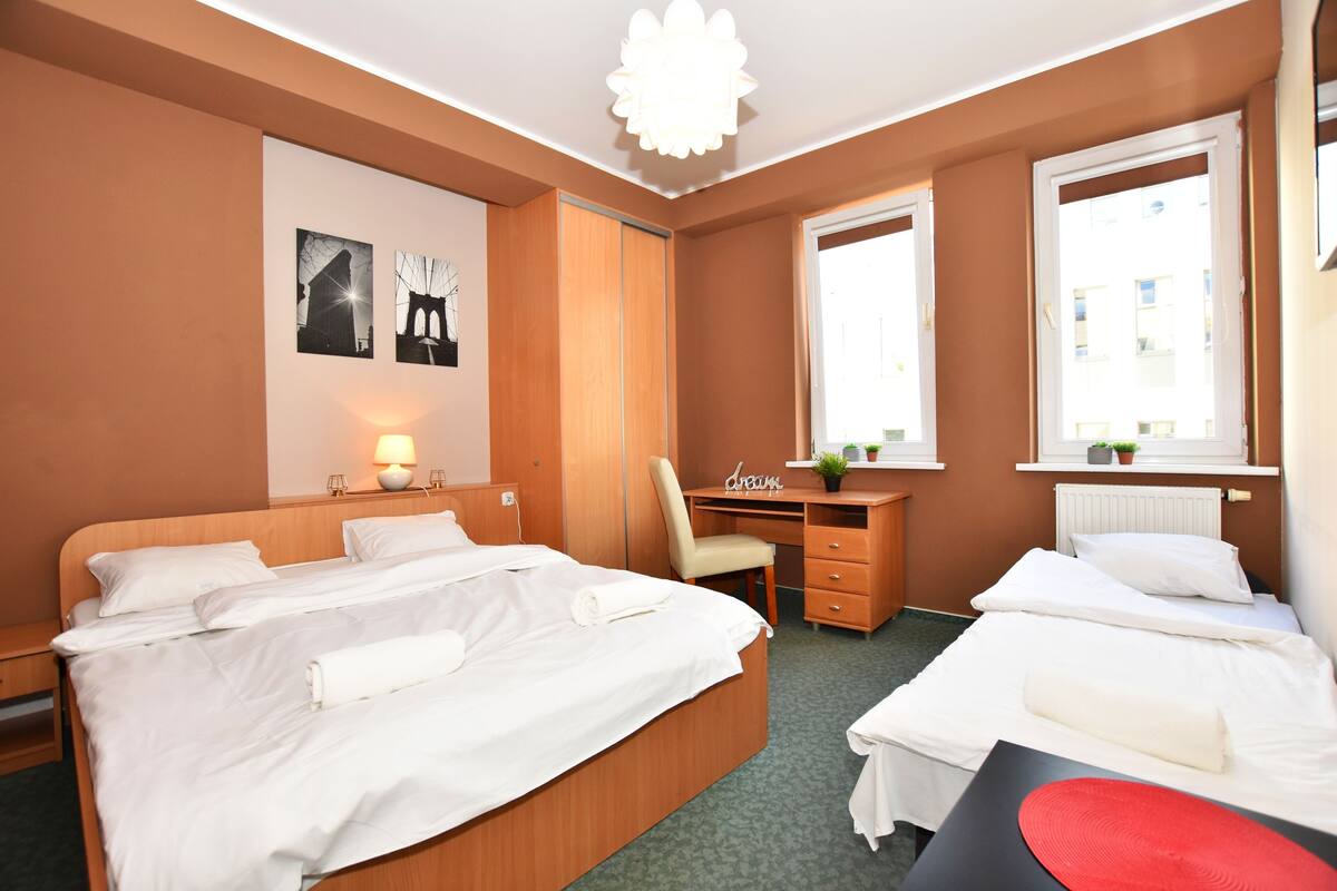 A hotel room with two beds, a desk, a chair, a wall-mounted lamp, and a window with a view of buildings.
