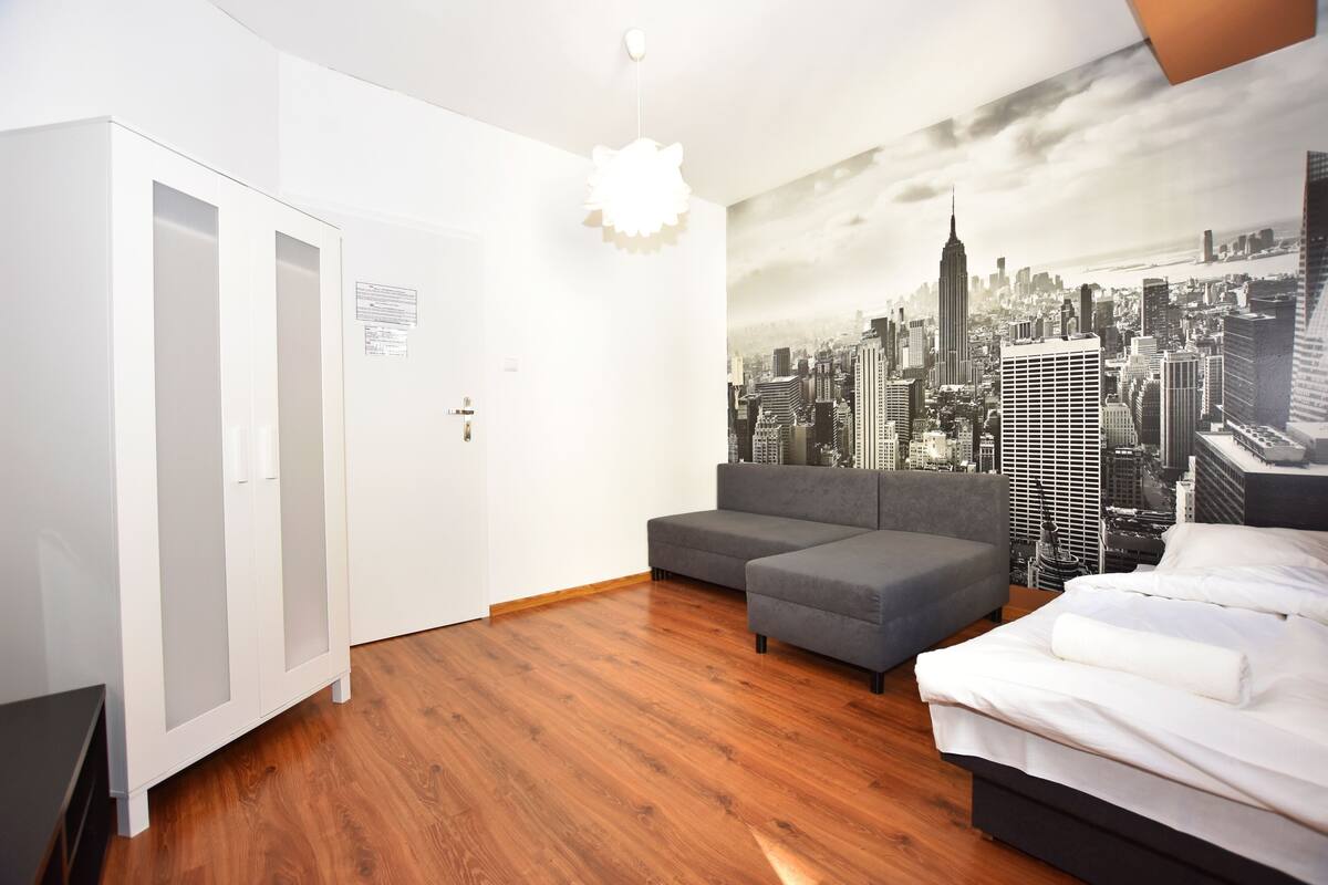 A bedroom with a gray sofa, a bed, and a cityscape mural on the wall.