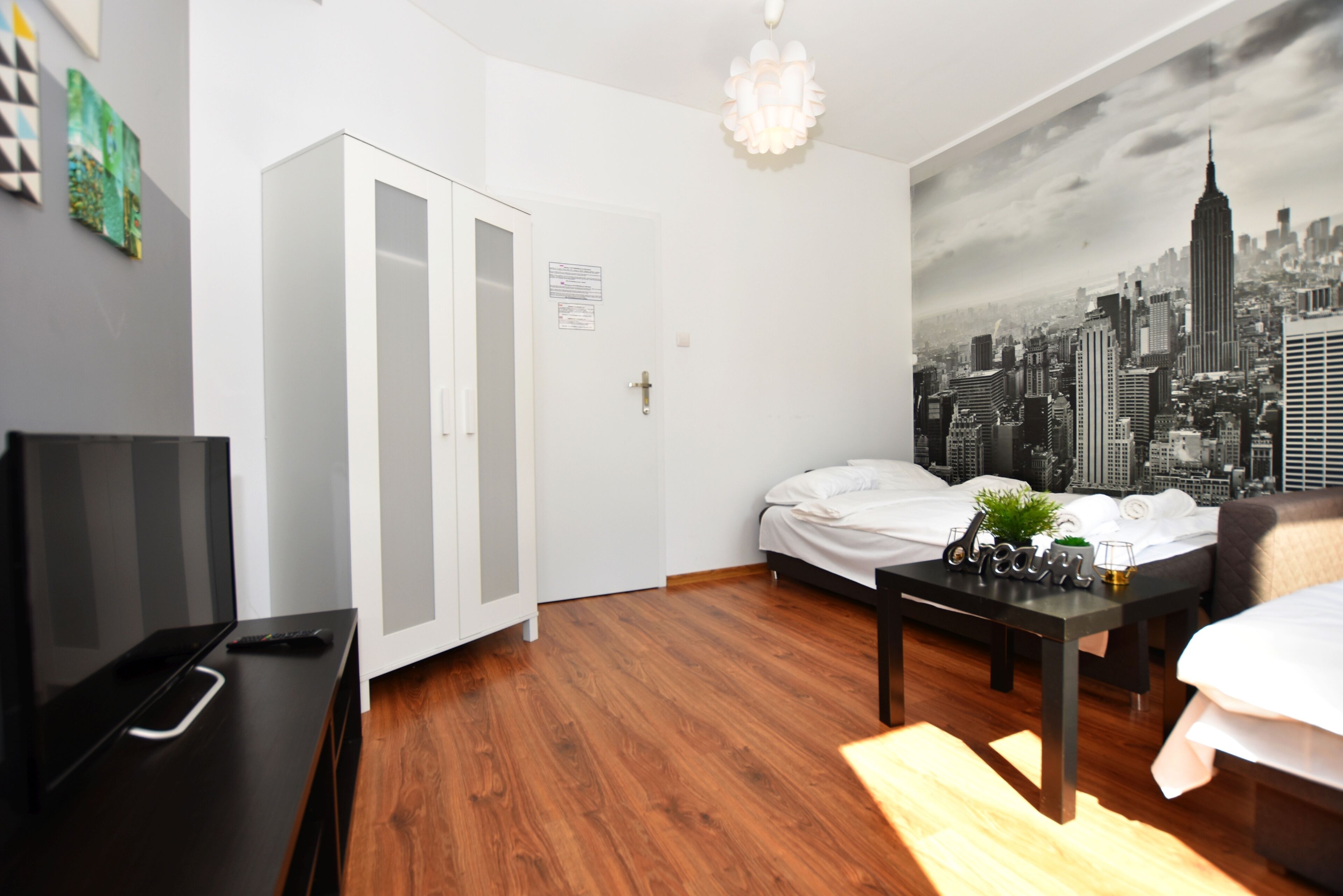 A modern hotel room with a wooden floor, a bed with white linens, a black coffee table, a TV, and a cityscape mural on the wall.