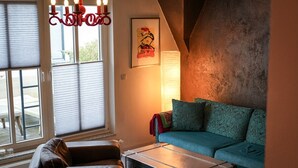 A living room with a sofa, a coffee table, a window with blinds, a chandelier, and a framed picture on the wall. - Apartment Cajus - centrally located "Am Alten Strom" with quiet roof terrace (Warnemünde)
