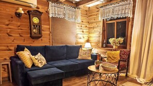 Interior acogedor de una cabaña de troncos con un sofá azul oscuro, piso de madera, una mesa redonda de metal con una lámpara y un ventanal con cortinas de encaje.