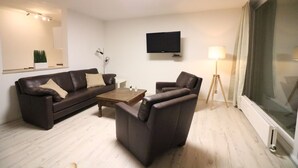Ein Wohnzimmer mit einem braunen Ledersofa, einem braunen Ledersessel, einem Holztisch, einem flachbildfernseher an der Wand, einer Stehlampe und einem großen Fenster mit Vorhängen.