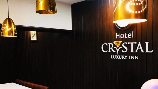 Un'area reception con un bancone bianco, tre lampade a sospensione e una ventola a soffitto. Le pareti sono rivestite in pannelli di legno scuro, e si distingue il logo di 'Hotel Crystal Luxury Inn'.