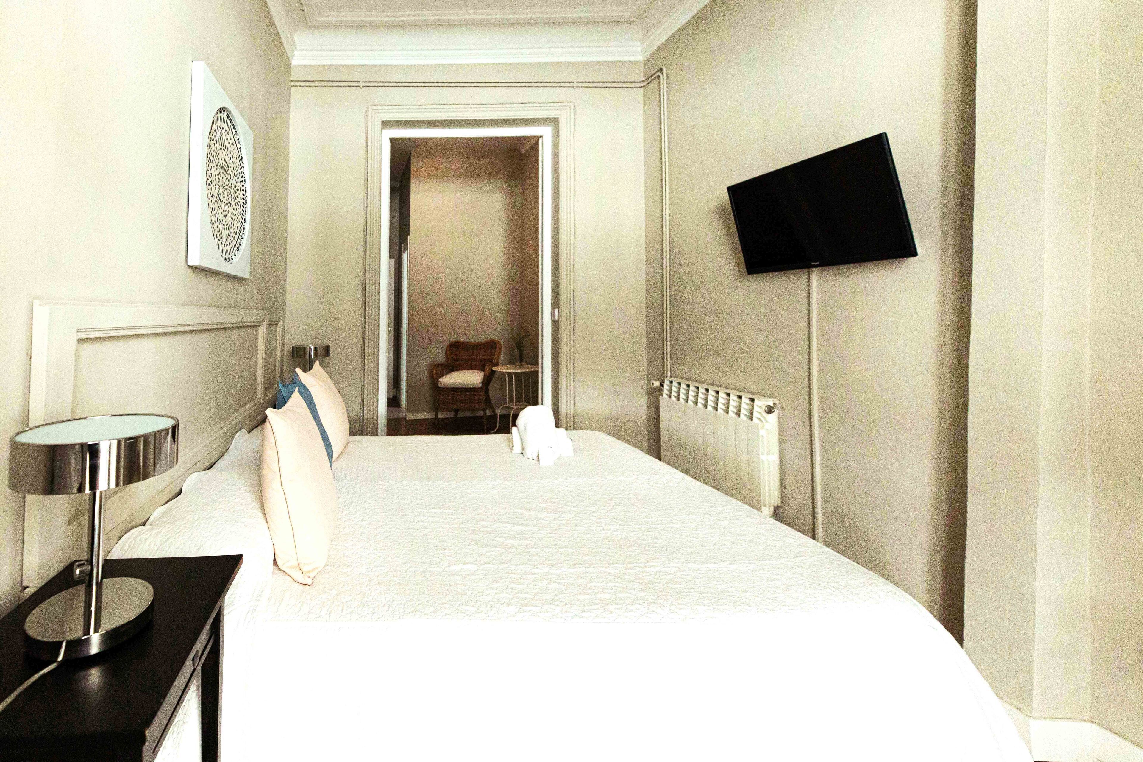 A hotel room with a large bed, a bedside table with a lamp, a flat-screen TV mounted on the wall, and a door leading to another room.