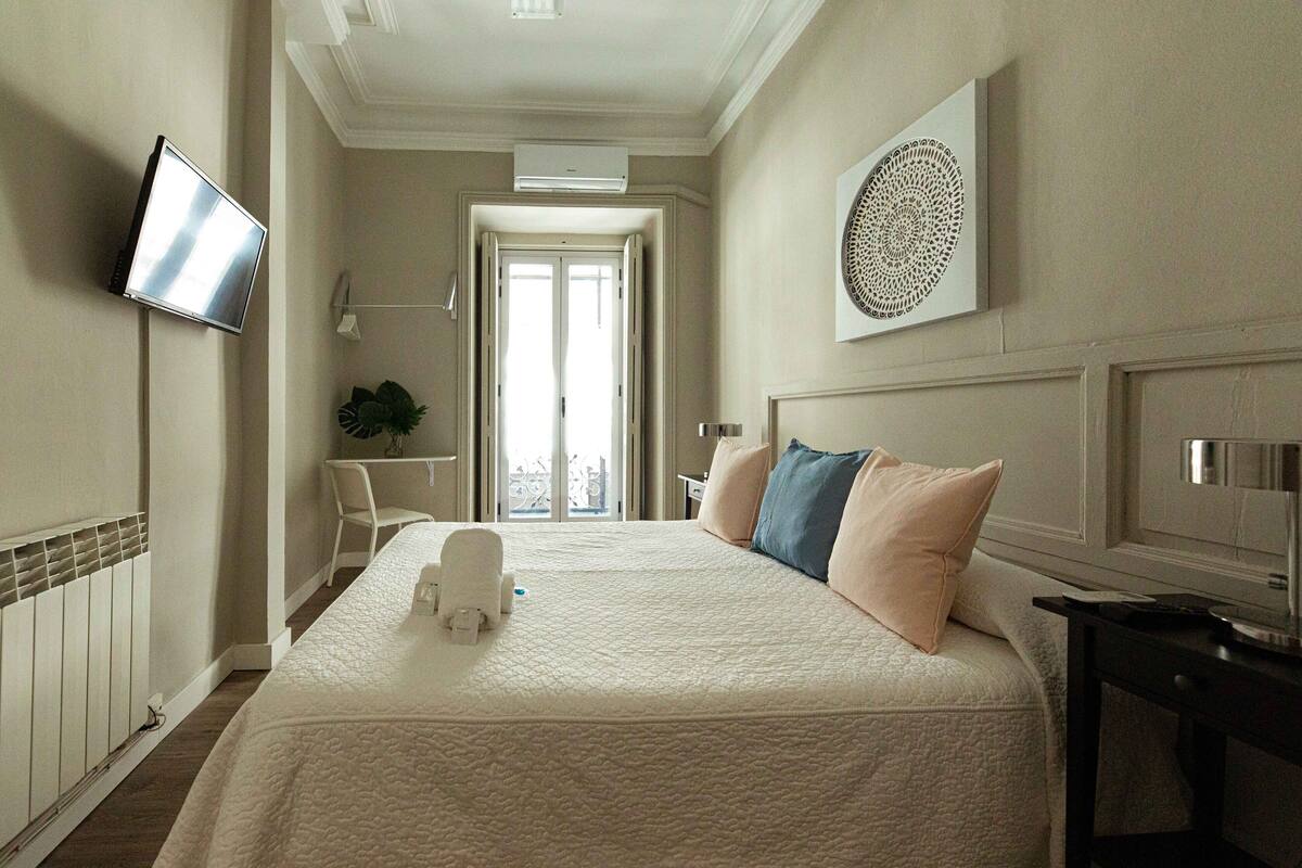 A hotel room with a large bed, a flat-screen TV, a decorative wall piece, and a view of the city through the door.