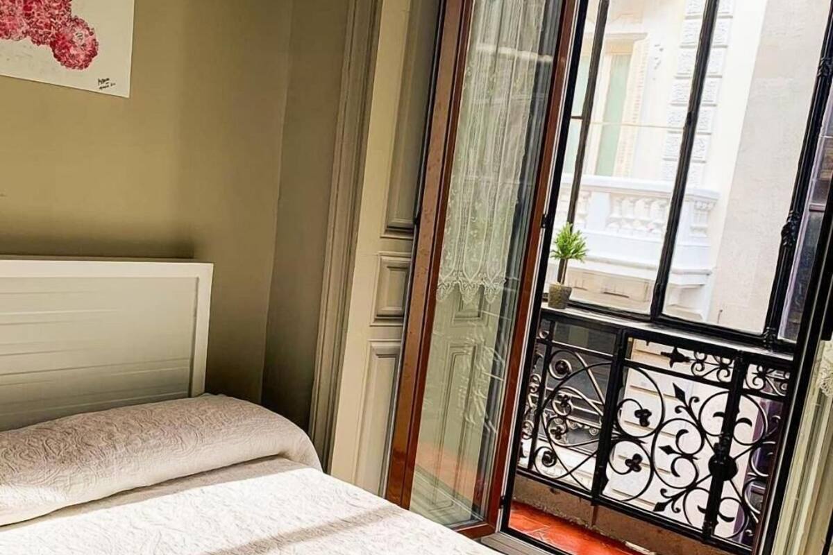 A hotel room with a bed, a window with a view of buildings, and a balcony with a railing.
