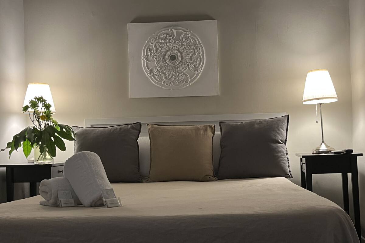 A neatly made bed with a white duvet, two pillows, and a folded towel on a headboard. There is a decorative wall piece above the bed and two bedside tables with lamps.