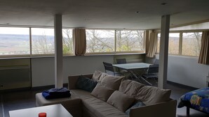 Una habitación con sofá, mesa de comedor y cama. Amplias ventanas con cortinas.