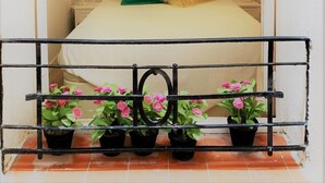 Ein Schlafzimmer mit einer ziegelmuster-ähnlichen Wand, einem Bett mit einem grünen Kissen und einer hängenden, gelben Leuchte. Vor einem Metallgeländer stehen Blumentöpfe mit rosa Blüten.
